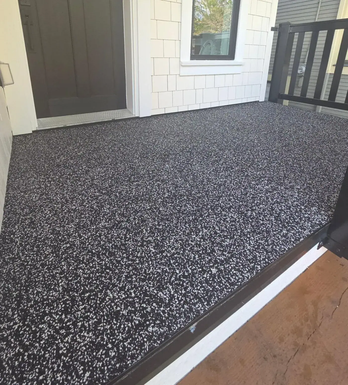 Front entry porch surfaced in black and white rubber granules, beside a dark front door and white shingle siding.