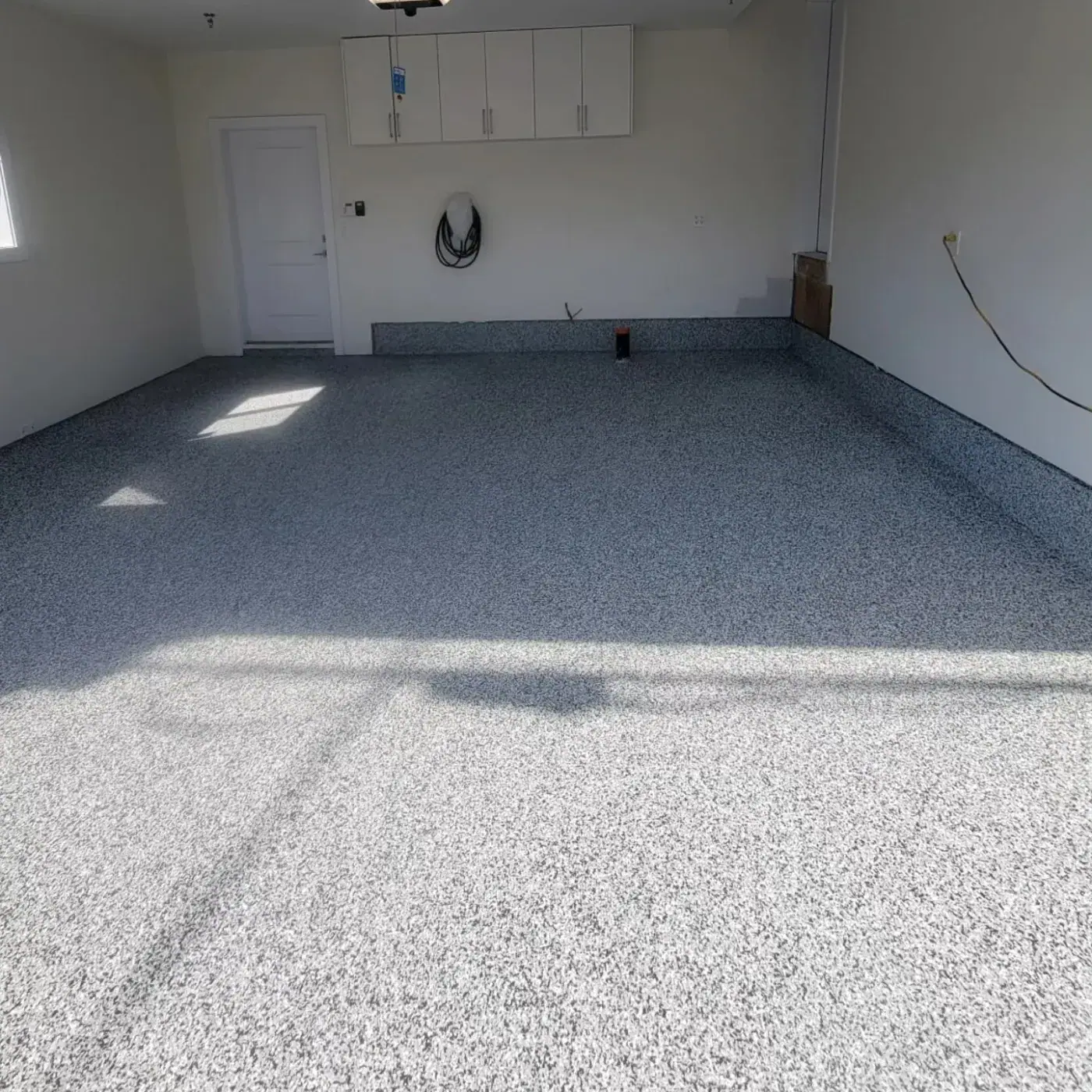 Bright white garage with overhead cabinets and a coiled cable on a wall hook, finished with a pale grey flake floor.