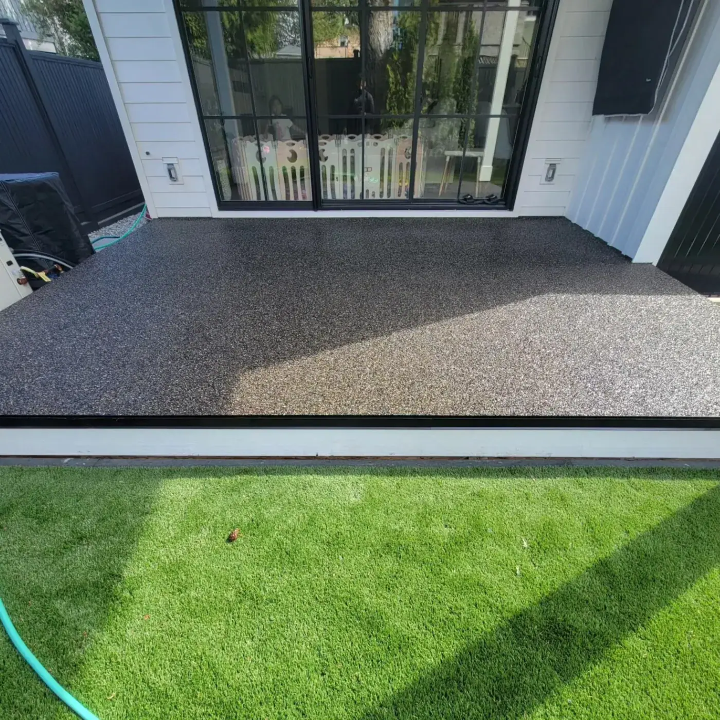 Rear patio in dark granular surfacing between black-framed glass doors and a strip of artificial turf.