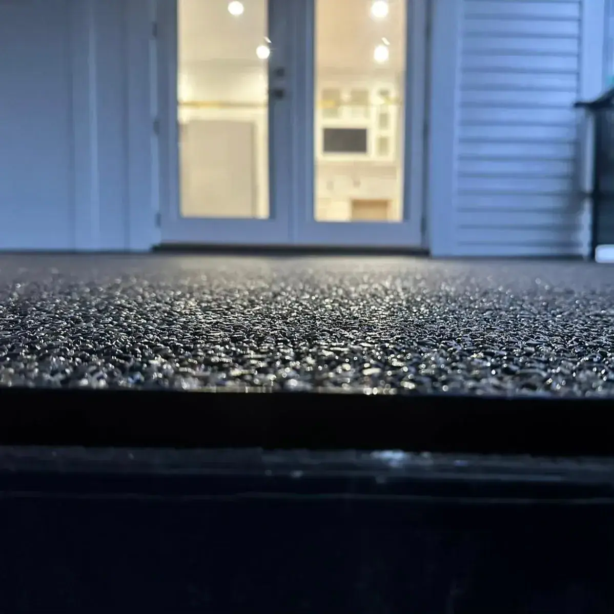 Night view along the edge of a dark granular patio surface, lit from the glass doors of the house behind.