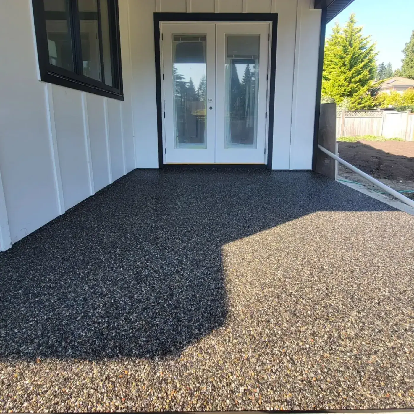 Covered patio at a white board-and-batten house, dark stone surfacing beneath French doors with afternoon shadow falling across it.
