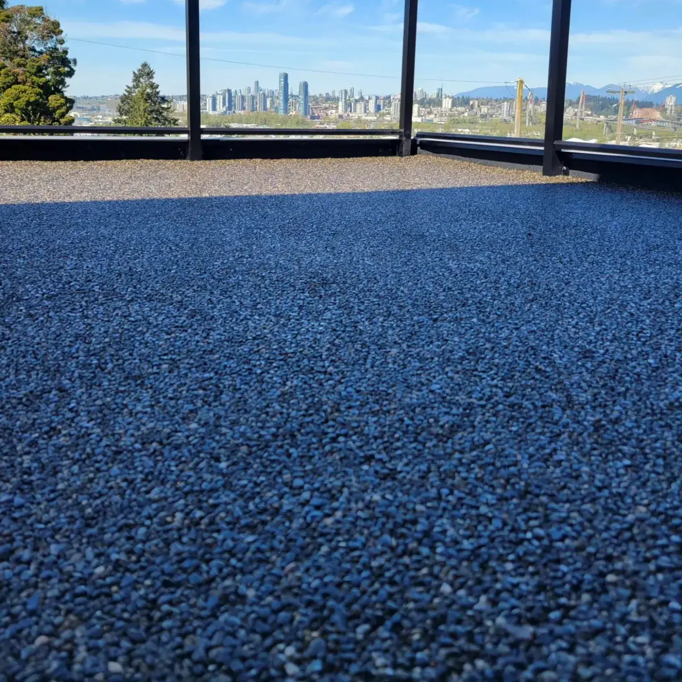 Roof deck coated in deep blue granular surfacing behind a glass railing, looking out over a city skyline.