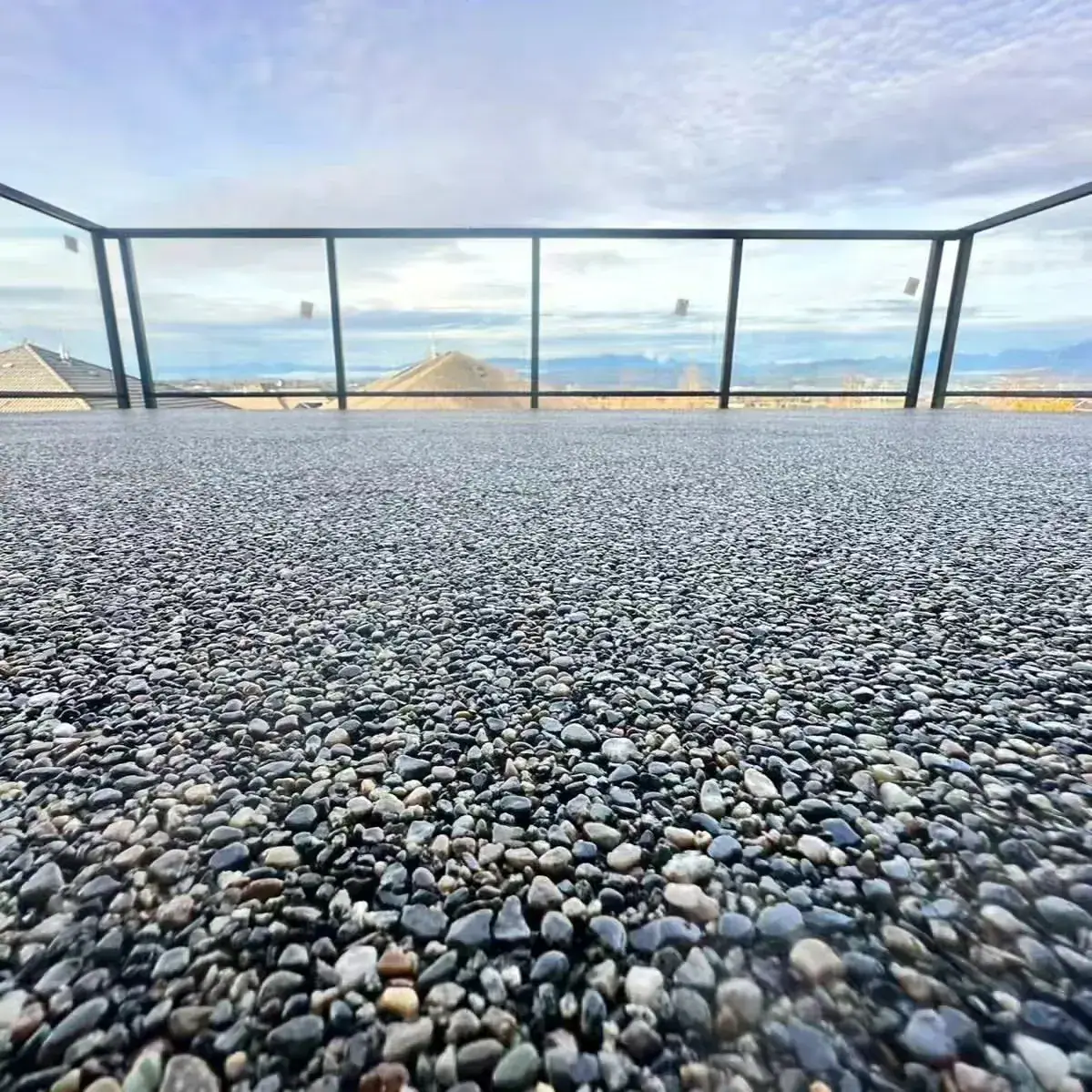 Rooftop deck finished in grey river-stone surfacing, with a black-framed glass railing and a view over rooftops to distant mountains.